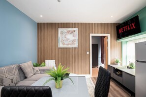 A modern living room with a sofa, a dining table with a plant, and a kitchen area with a refrigerator and a wall-mounted television displaying 'NETFLIX'. - Contemporary Kelham 1-Bed Pet-Friendly Apartment (Sheffield)