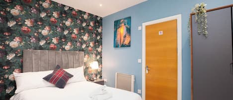 Une chambre d’hôtel avec un lit, une table de chevet, un mur recouvert d’un papier peint à motifs floraux, un tableau, une porte et une plante.