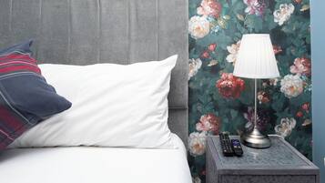 Uma cama com travesseiro branco e travesseiro xadrez, mesa de cabeceira com abajur e controles remotos, e papel de parede floral.