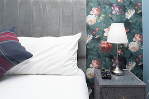 A bed with a white pillow and a plaid pillow, a bedside table with a lamp and remote controls, and a floral wallpaper.