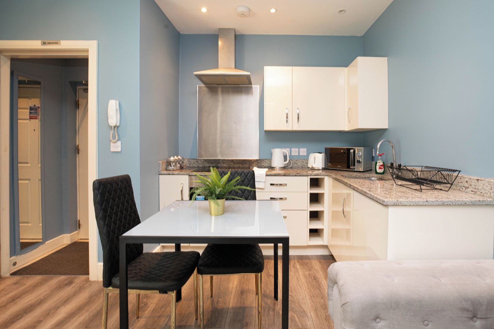 A compact kitchen with a dining area, including a table with two chairs, a microwave, a kettle, and a sink with a dish rack.