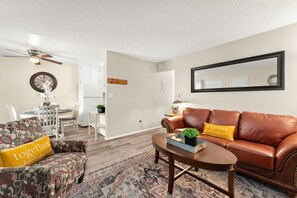A living room with a brown leather sofa, a patterned armchair, a coffee table, a dining area with a white table and chairs, and a ceiling fan.