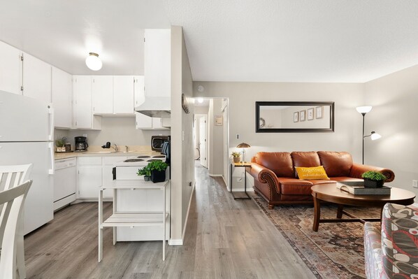 A modern kitchen with white cabinets, a dining area with a white table and chairs, a brown leather sofa, and a patterned rug.