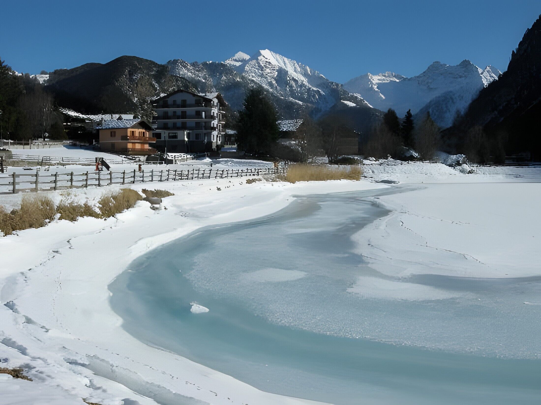 Paisaje nevado con un lago helado, montañas al fondo y un edificio de varios pisos.