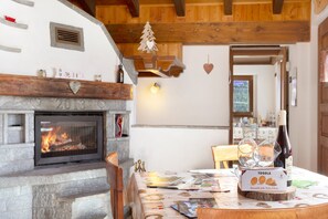Une pièce chaleureuse avec une cheminée, des poutres apparentes, une table à manger sur laquelle se trouve une boîte de Tegolet et une bouteille de vin.