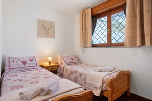 A bedroom with two single beds, a wooden headboard, a window with curtains, a wall-mounted lamp, and a deer head artwork.