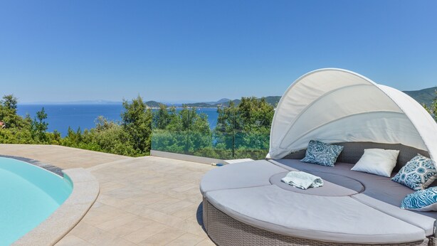 A poolside area with a view of the sea, a covered seating area with cushions, and a clear sky.
