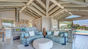A modern living room with a sectional sofa, a white ottoman, a kitchen area, and a view of an outdoor pool.