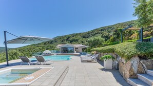 A pool area with lounge chairs, a hot tub, and a view of a hillside.