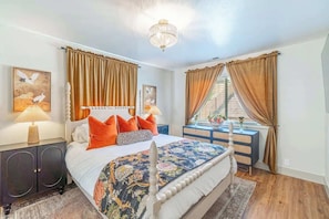 Un dormitorio con cama, cortinas, una lámpara de araña, cómoda y obras de arte en las paredes.