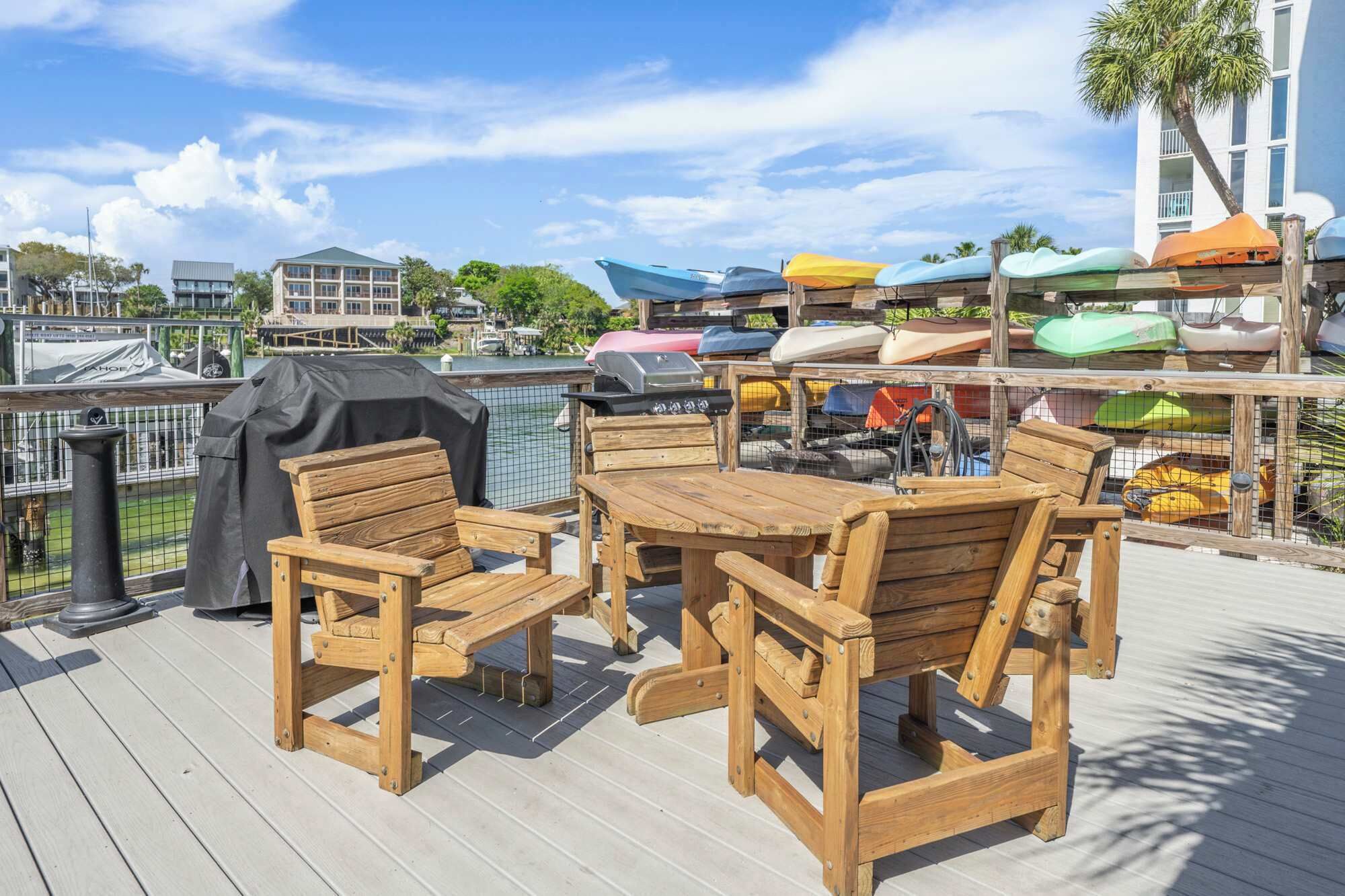 Une terrasse en bois avec une table et des chaises, un barbecue, et des kayaks colorés en arrière-plan.