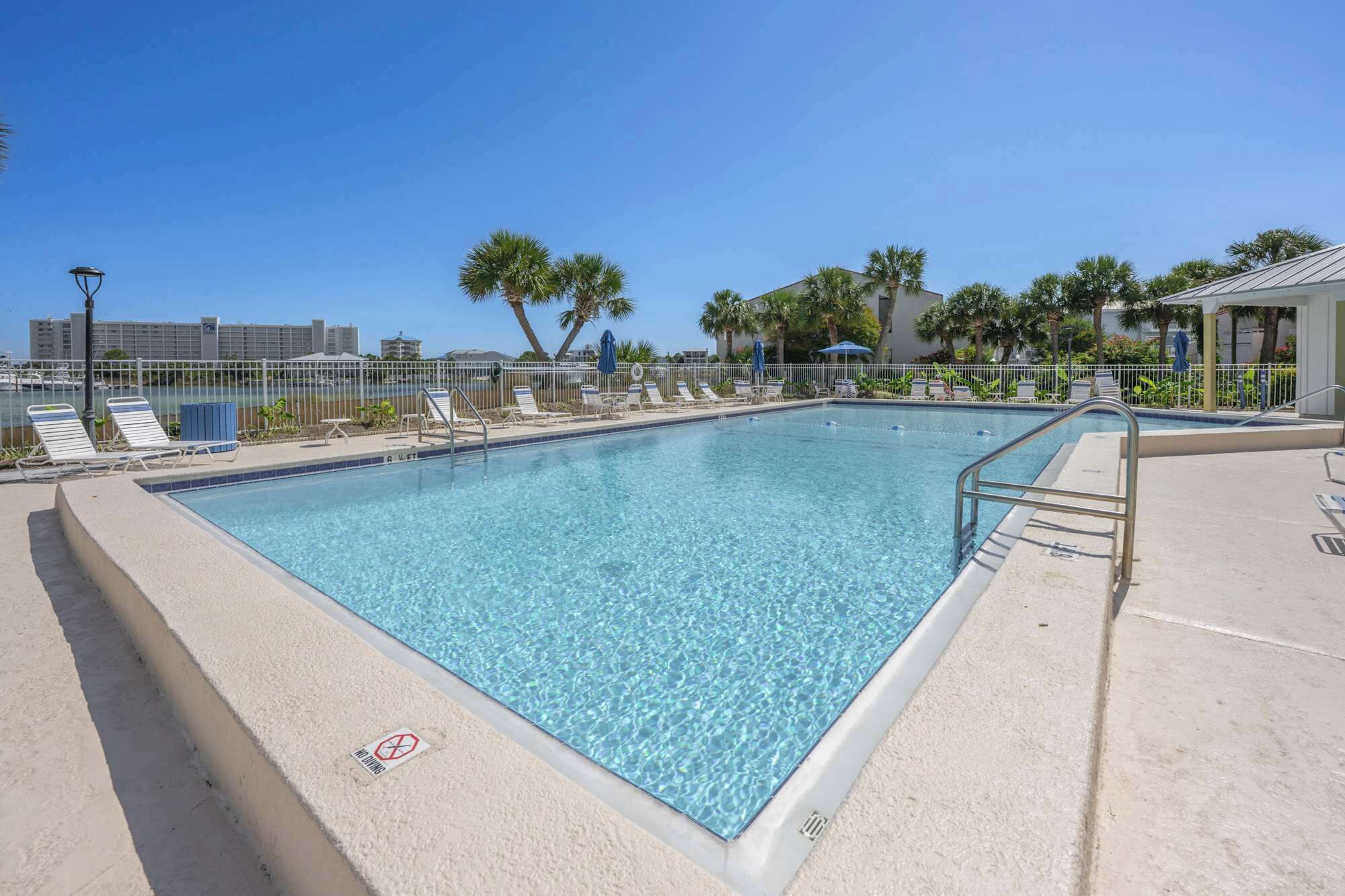A rectangular swimming pool with clear blue water, surrounded by a white fence and palm trees.