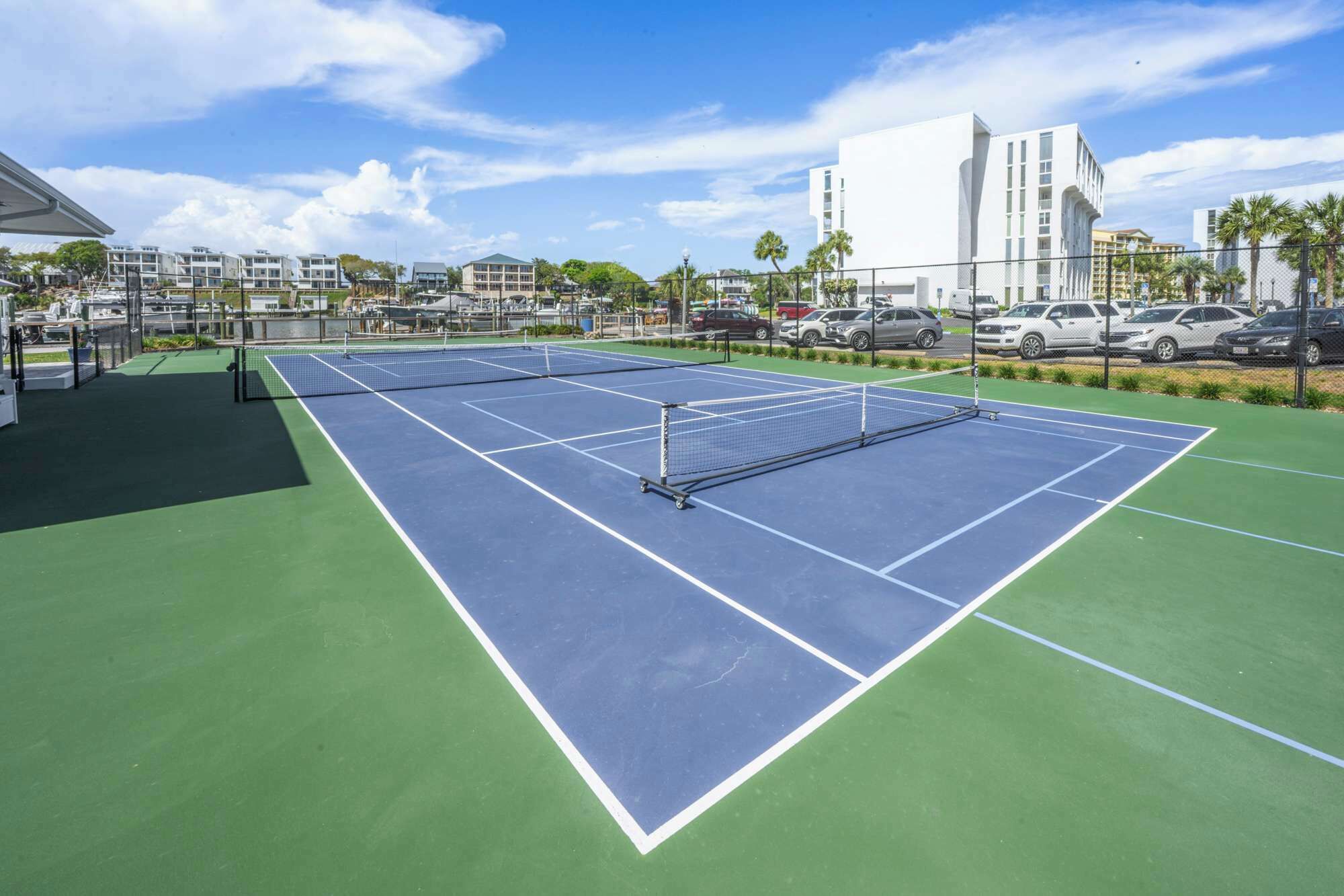 A tennis court with a net, surrounded by a fence, and adjacent to a parking area with several cars.
