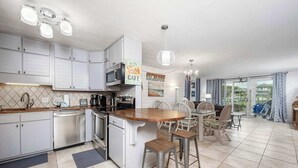 A modern kitchen with white cabinets, a central island, and bar stools. There is a dining area with a table and chairs, and a view of the ocean through large windows. - Destin Retreat: Walk to Private Beach & Pool (Destin)