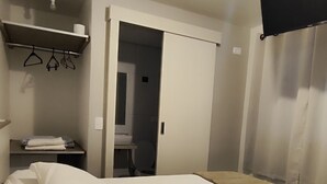 Quarto de hotel com uma cama, uma escrivaninha com controle remoto, uma estante com cabides, um vaso sanitário e uma porta que dá para outro quarto.