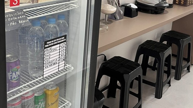 Geladeira com sanduíches, frutas e bebidas. Há também banquetas e uma cafeteira no balcão.