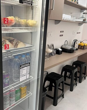 Geladeira com sanduíches, frutas e bebidas. Há também banquetas e uma cafeteira no balcão.