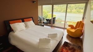 Una habitación de hotel moderna con una cama grande, un sillón amarillo y un balcón con vistas a zonas verdes y un lago.