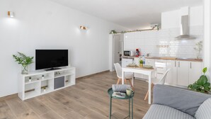Un soggiorno moderno con una TV a schermo piatto, un divano, un tavolo da pranzo con sedie e un angolo cottura con mobili bianchi e un forno a microonde.