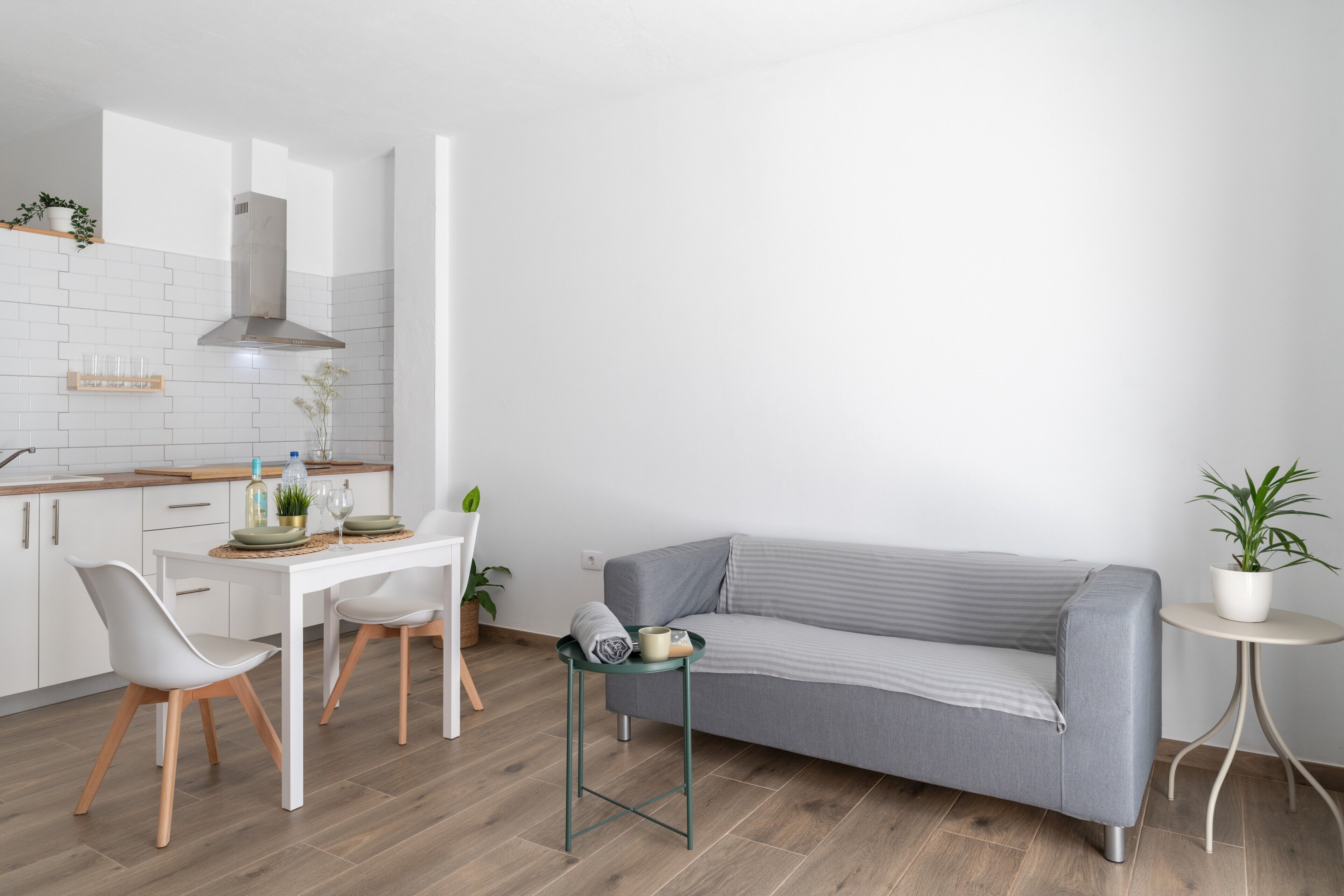 Eine moderne Küche mit weißen Schränken, einer Holz Arbeitsplatte, einer Edelstahl-Dunstabzugshaube, einem Esstisch mit zwei Stühlen, einem grauen Sofa und einem kleinen runden Tisch mit einer Pflanze.