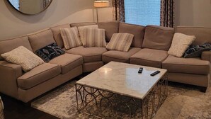 Een moderne woonkamer met een hoekbank, een ronde spiegel, een vloerlamp en een groot raam met gordijnen.