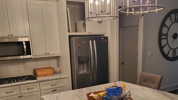 Een keuken met witte kasten, een roestvrijstalen koelkast, een inbouwoven, een marmeren eiland met een mand snacks, en een grote wandklok.