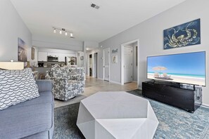 A modern living room with a sofa, a coffee table, a flat-screen TV, and a kitchen in the background. - Sunshine & Whiskey Condo for 6 With Resort Amenities Near Grand Boulevard (Miramar Beach)