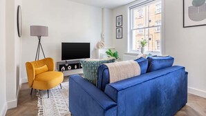 A living room with a blue sofa, a yellow armchair, a flat-screen TV, and a large window.