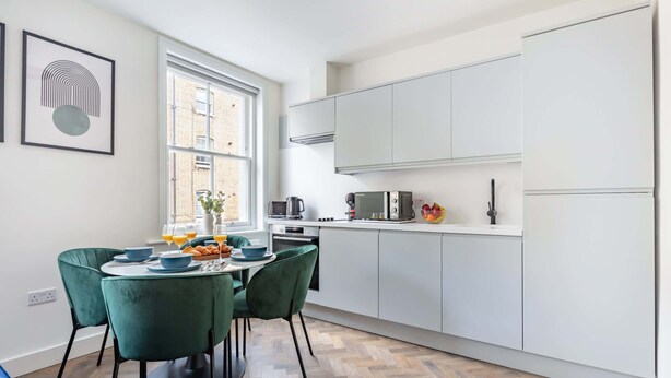 A modern kitchen with white cabinets, a dining table with green chairs, and a window with a view of buildings.