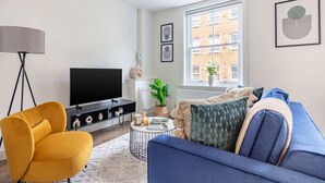 A modern living room with a blue sofa, a mustard yellow armchair, a flat-screen TV, a floor lamp, and a large window with a view of buildings.