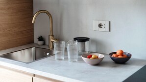 Una cocina moderna con un lavamanos de acero inoxidable, un grifo dorado y una encimera de mármol. Hay dos recipientes de vidrio y un bol con frutas sobre la encimera.