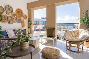 A modern living room with a sofa, round table, and wicker furniture. There are plants and decorative items on the walls. - Sundrop 1-Bedroom Apartment in Larnaca (Larnaca)