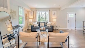 A spacious living room with a dark leather sofa, two armchairs, a glass coffee table, and a large window with curtains. - Walk to the stadium! - Lighthouse on Glenwood (Annapolis)