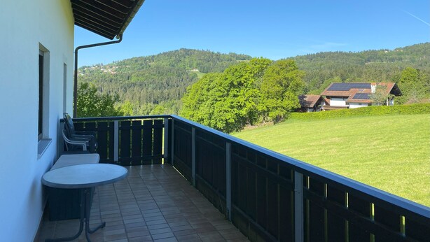 Ein Balkon mit Tisch und Stühlen, Blick auf ein grünes Feld und ein Haus mit Solaranlage, sowie ein klarer, blauer Himmel.
