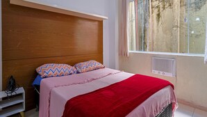 Un dormitorio con cabecera de madera, una cama con manta roja, dos almohadas estampadas, una ventana con vistas a árboles y un aire acondicionado de pared.