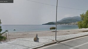 Uma estrada costeira com uma praia de pedras, uma estátua e uma montanha ao fundo.