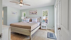 Une chambre à coucher comprenant un lit, des tables de chevet, un ventilateur de plafond et une fenêtre avec des stores.