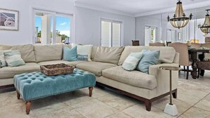 Ein geräumiges Wohnzimmer mit einem beigefarbenen Ecksofa, einem blauen Hocker und einer Pendelleuchte.