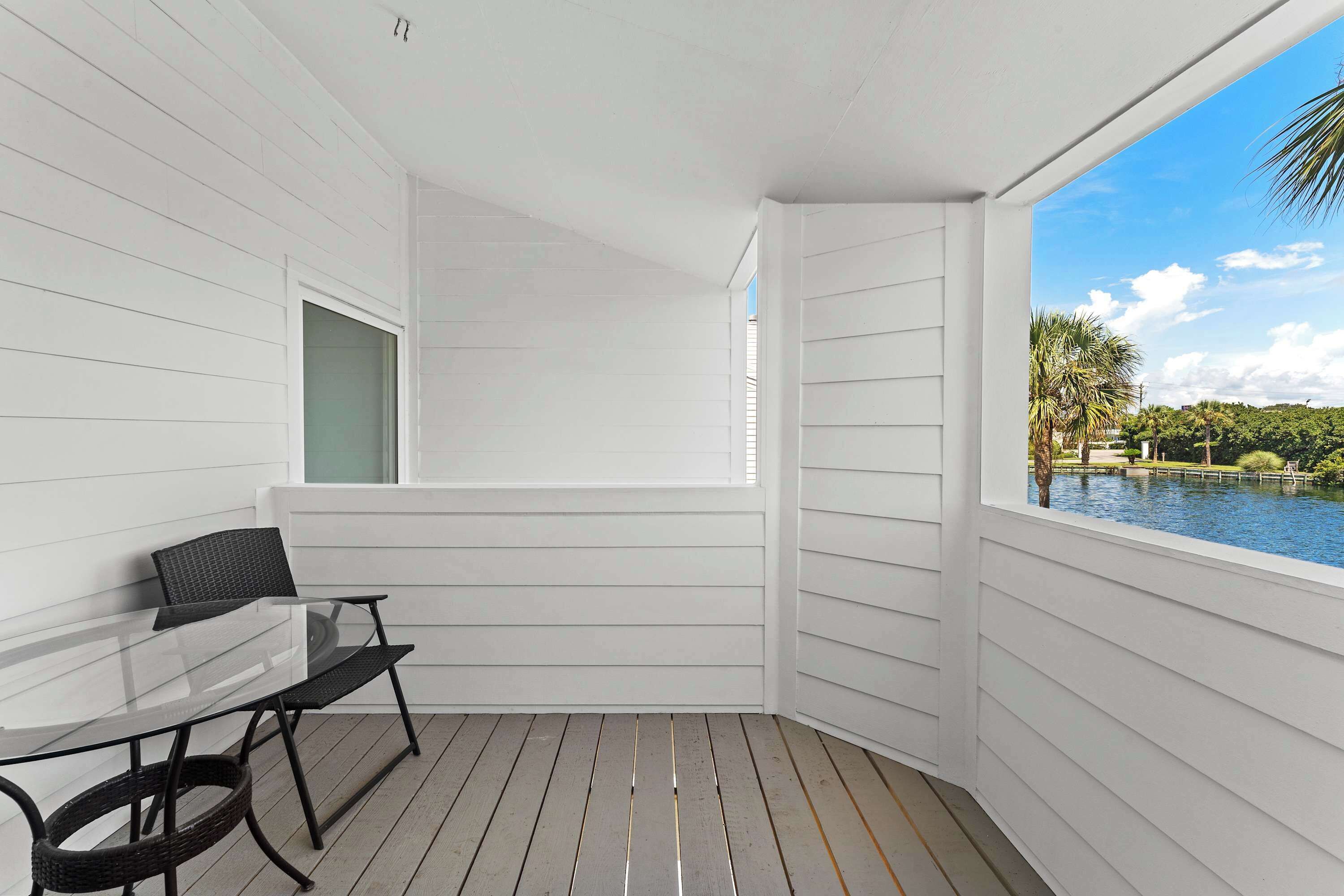 A balcony with a glass table and chairs, white siding walls, and a view of a lake with trees.