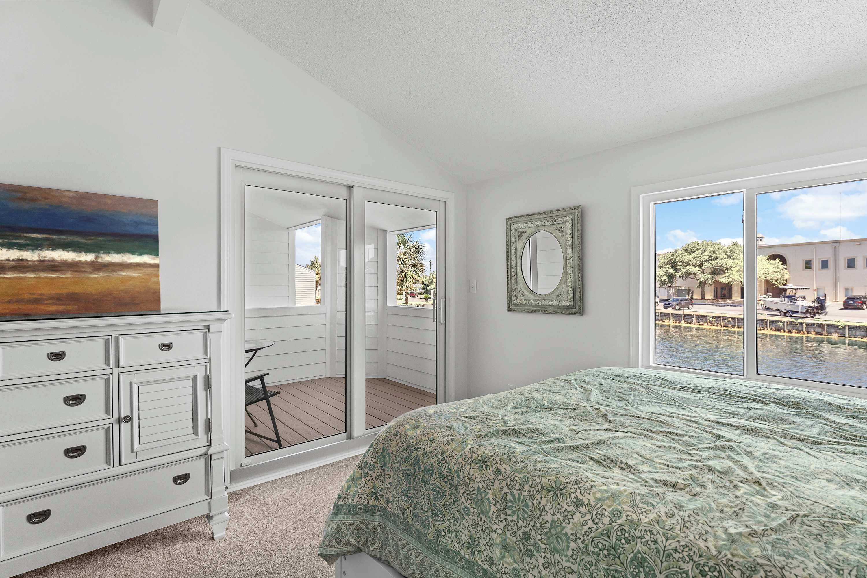 A bedroom with a bed, a dresser, a painting, a mirror, and a view of a balcony and water.