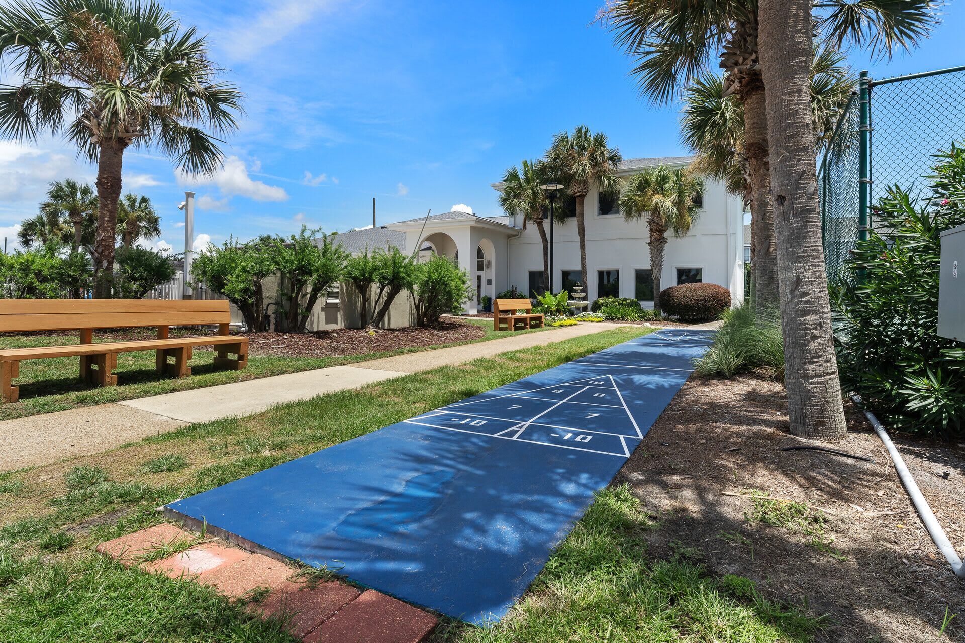 A blue shuffleboard court with a wooden bench and palm trees in a landscaped area.