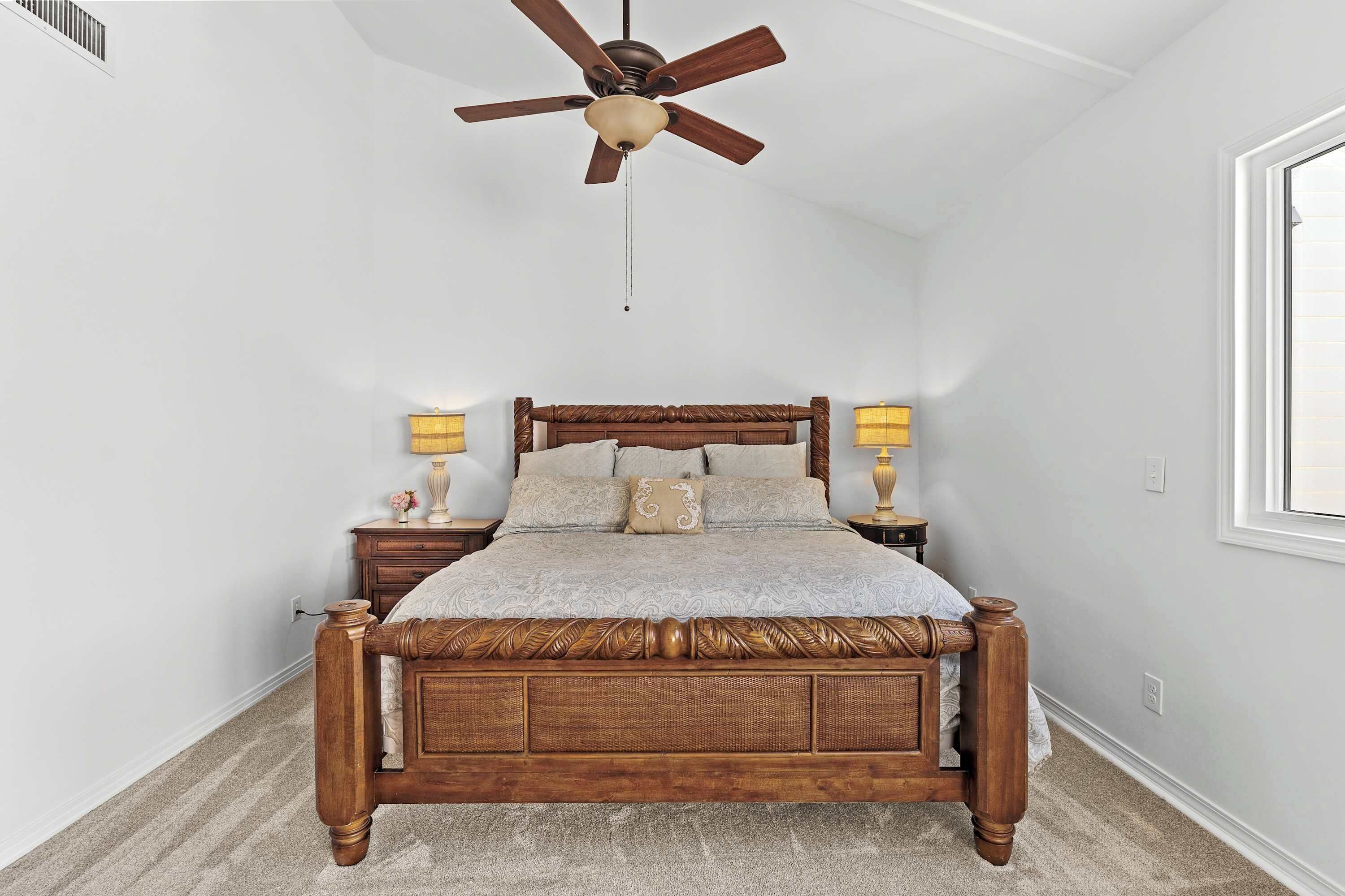 A bedroom with a wooden four-poster bed, two bedside lamps, a ceiling fan, and a large window on the right.