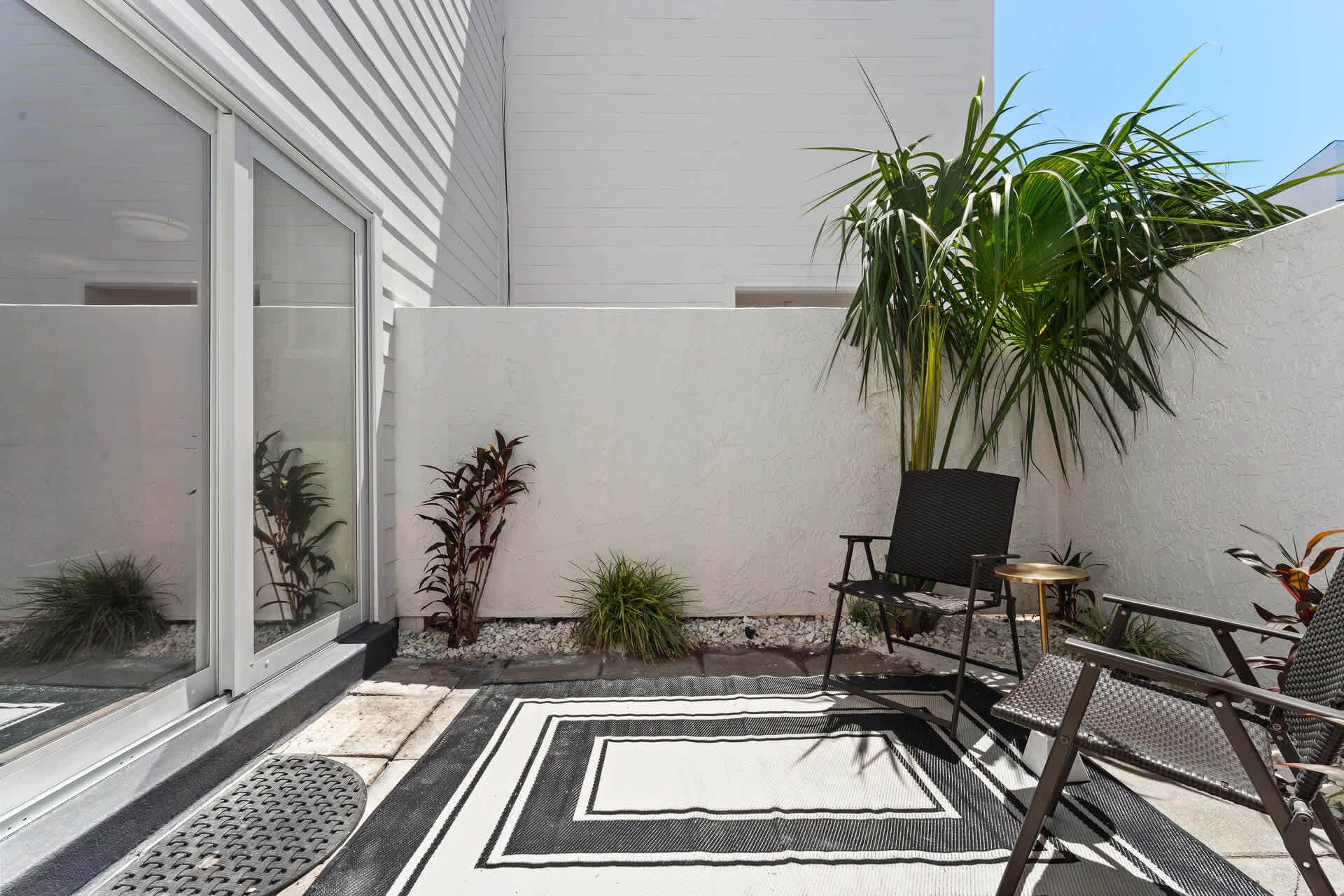 A small outdoor patio area with a black and white striped rug, a black chair, and a tall green plant.