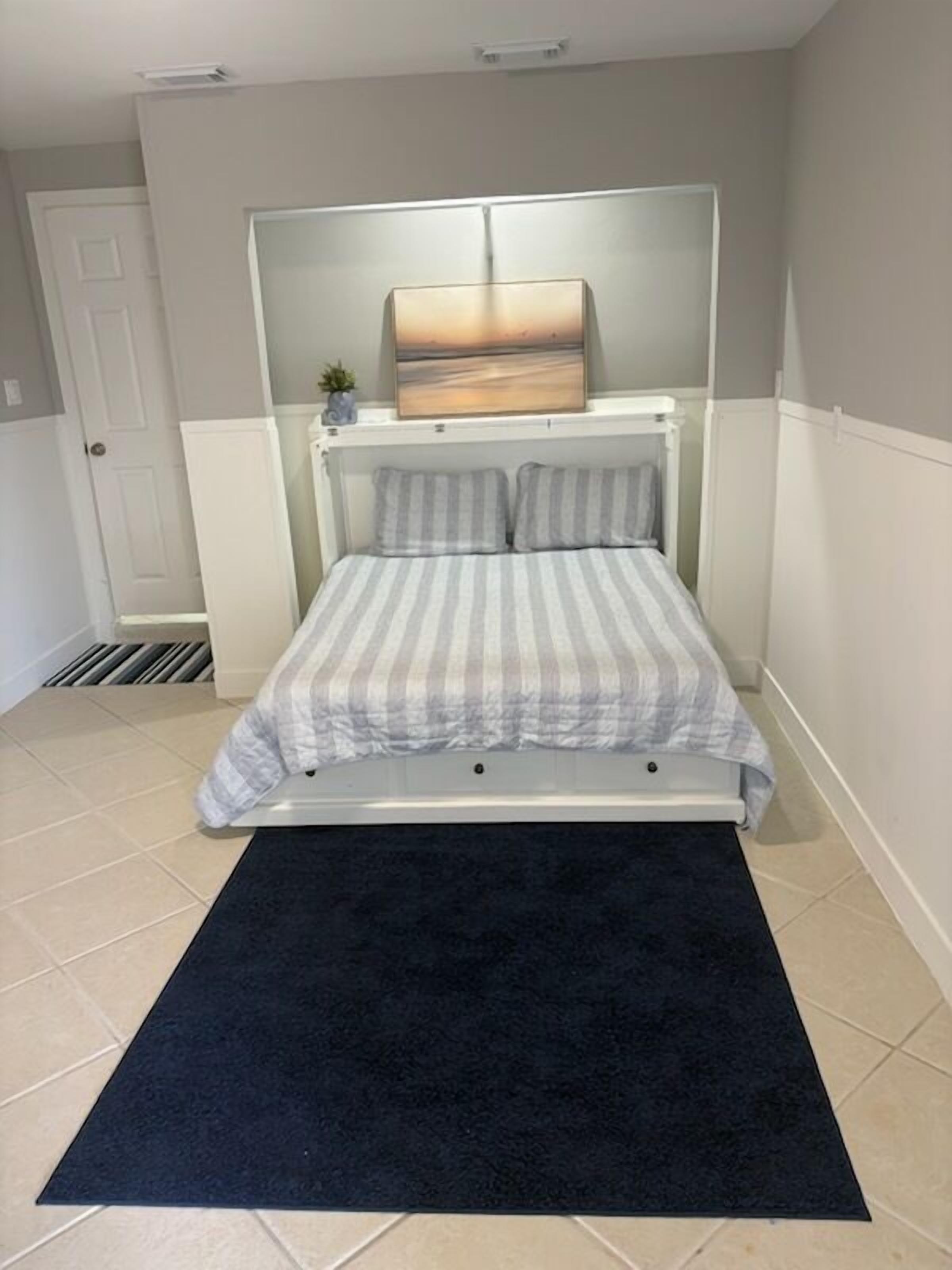 Een slaapkamer met een bed, een donker vloerkleed, een schilderij aan de muur en een kleine plant op het nachtkastje.