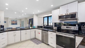 A modern kitchen with white cabinets, stainless steel appliances, and a large window providing a view of the beach.