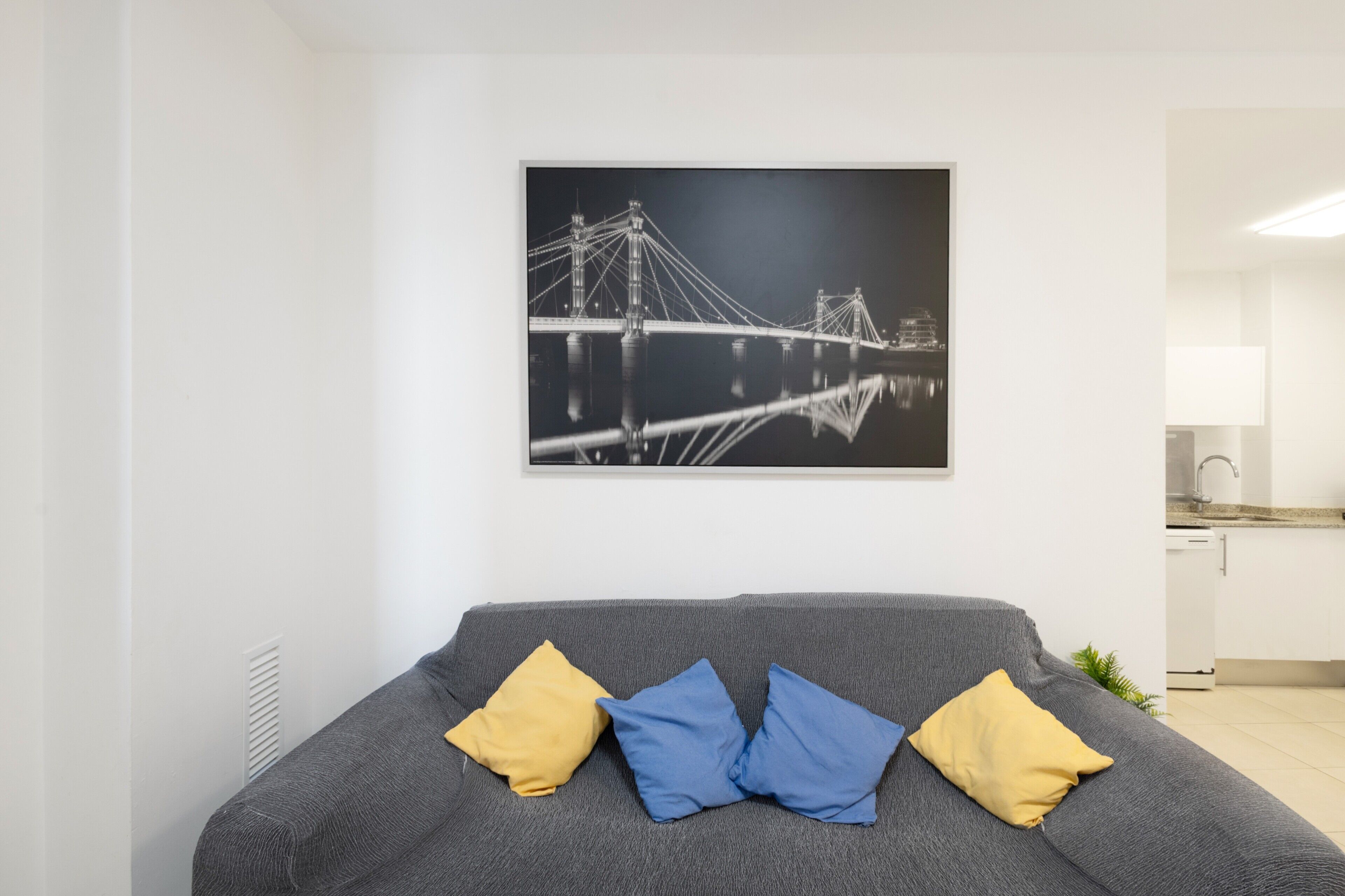 A modern living room with a gray sofa, a black and white photograph of a bridge, and a kitchen in the background.