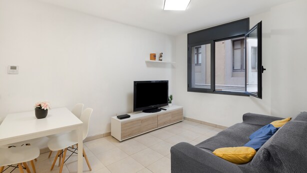 Ein modernes Wohnzimmer mit Flachbildfernseher, einer Couch mit Kissen, einem Esstisch mit Stühlen und einem Fenster mit Blick ins Freie.