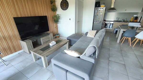 A modern living room with a flat-screen TV, grey sofa, wooden coffee table, and a kitchen area in the background.