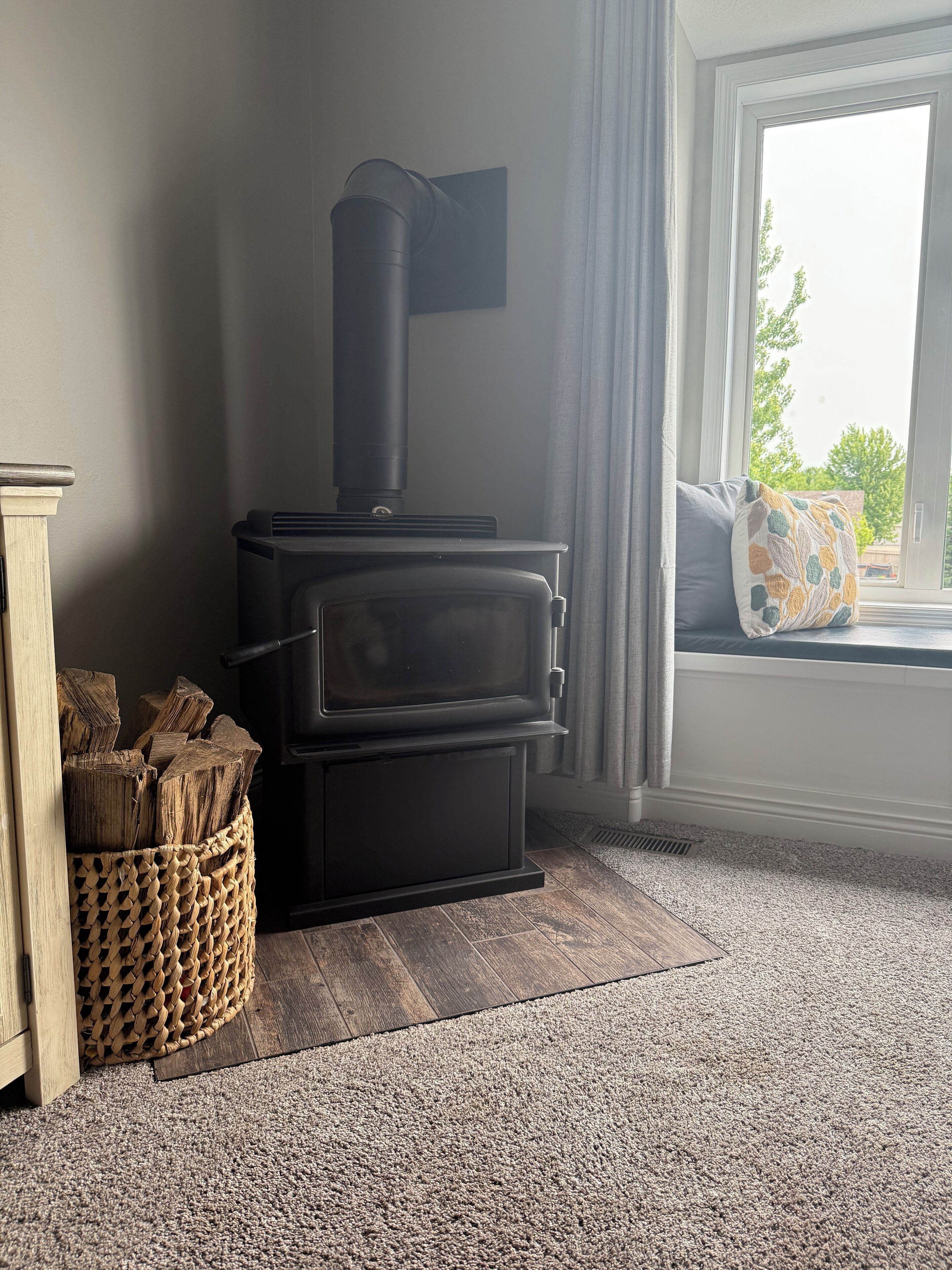 A wood-burning stove in a corner of a room with a window, a woven basket of firewood, and a grey carpet.