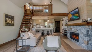 A spacious living area with a high ceiling, a stone fireplace, a kitchen with a central island, and a staircase leading to an upper level.